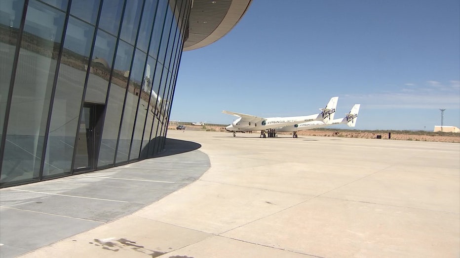 A photo showing a Virgin Galactic spacecraft outside, to the right o a glass-paned building