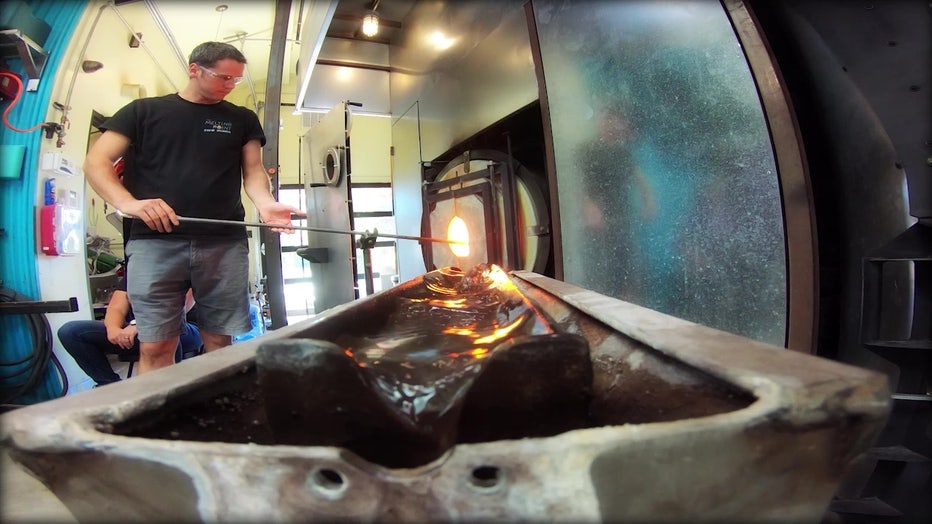 Photo of a man holding a pole containing molten glass, in front of a glass furnace