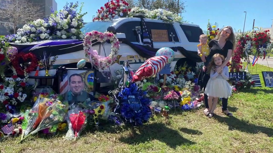 Deputy Michel Magli's family stands next to a patrol vehicle surrounded by flowers honor the deputy after he was killed in the line of duty. 