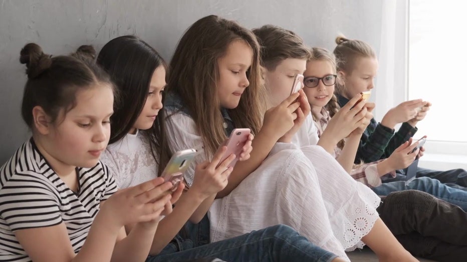 A group of children sit side-by-side looking at cell phones. 
