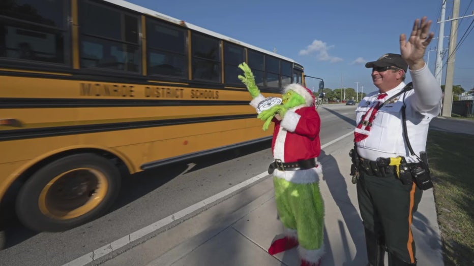 In this photo provided by the Florida Keys News Bureau, Monroe County Sheriff's Office Colonel Lou Caputo, left, costumed as the Grinch, and Deputy Andrew Leird, right, wave at a school bus rolling on the Florida Keys Overseas Highway Tuesday, Dec. 13, 2022, in Marathon, Fla. When drivers are pulled over for slightly speeding through a school zone, Caputo offers them the choice between an onion or a traffic citation. It's a holiday tradition in the Keys that Caputo began 20 years ago to educate drivers that Keys schools remain in session through Dec. 16.