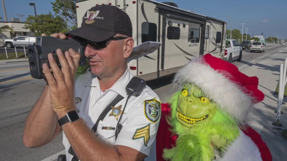In this photo provided by the Florida Keys News Bureau, Monroe County Sheriff's Office Colonel Lou Caputo, right, costumed as the Grinch, watches Sgt. Greg Korzan, left, as he uses a laser speed detector to check speeds of motorists traveling through a school zone on the Florida Keys Overseas Highway Tuesday, Dec. 13, 2022, in Marathon, Fla. For drivers slightly speeding through the area, Caputo offers them the choice between an onion or a traffic citation.