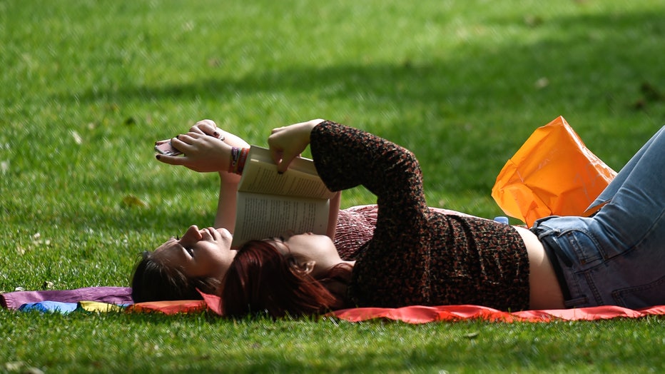 A woman reads a book next to her friend who is on her phone while relaxing in the sunshine in this file photo taken on May 15, 2019 in London, England.