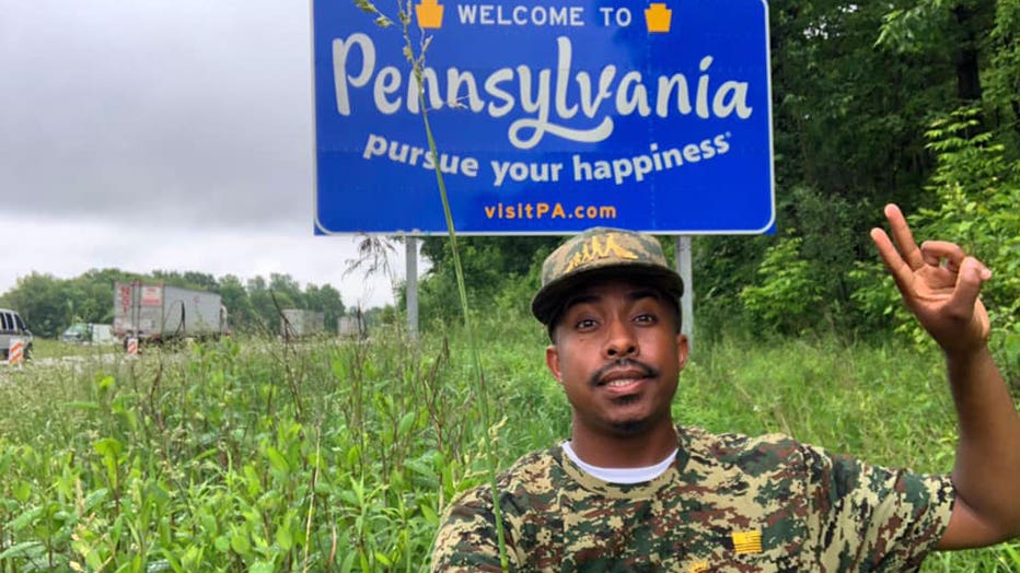 Rodney Smith Jr., a young black man, smiles in front of the Welcome to Pennsylvania sign on the state border. He is wearing a camouflage-colored hat and shirt.