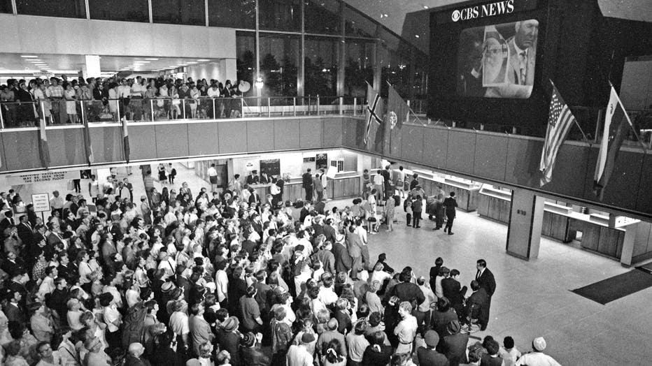 As Old Glory hangs over them, thousands of passengers and workers at Kennedy Airport International Arrivals Building stand or sit in awe as they watch on giant TV screen another American flag being raised on the moon. The crowd stands three deep on the balcony of building. Earlier, as the Eagle touched down on the moon's Sea of Tranquility, the atmosphere at the airport was anything but tranquil as the thousands whooped and cheered for their heroes.