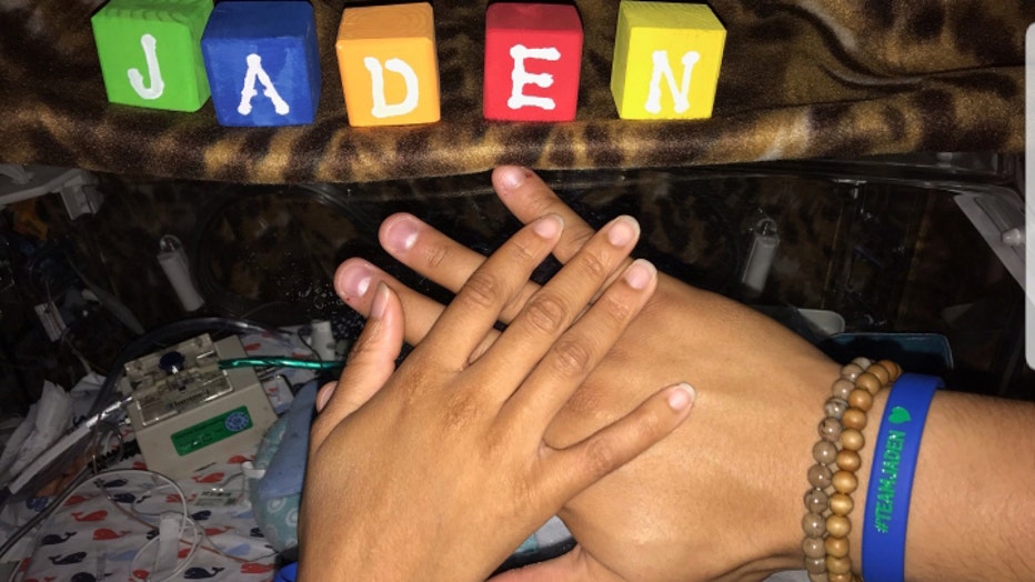 Jaden's parents hold their hands above his incubator, displaying bracelets that read