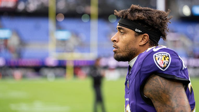Jaire Alexander in a Baltimore Ravens Uniform, without a helmet, looking to the left, with a football field blurred in the background.