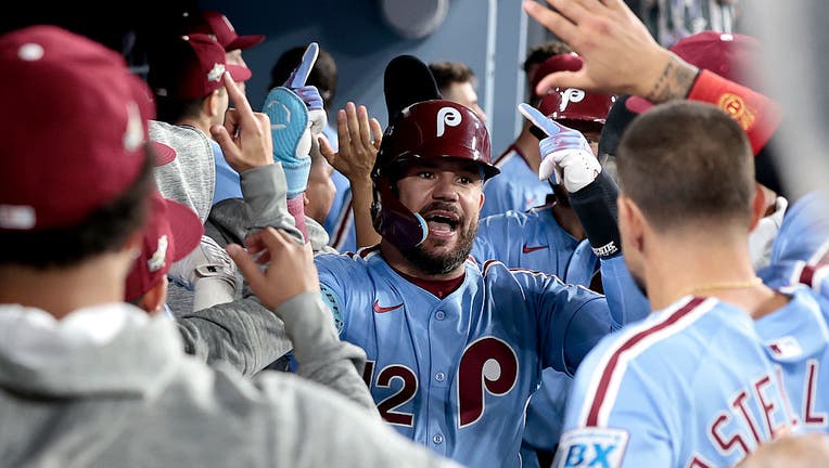 Philadelphia Phillies OF Kyle Schwarber celebrating in the dugout in the baby blue Phillies uniform and maroon batting helmet, with his hands in the air.
