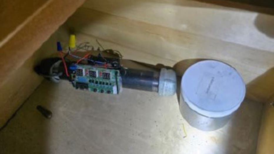 A homemade pipe bomb, with a circuit board, wires and a black pipe with silver metal caps, sitting in a box next to a metal cannister.