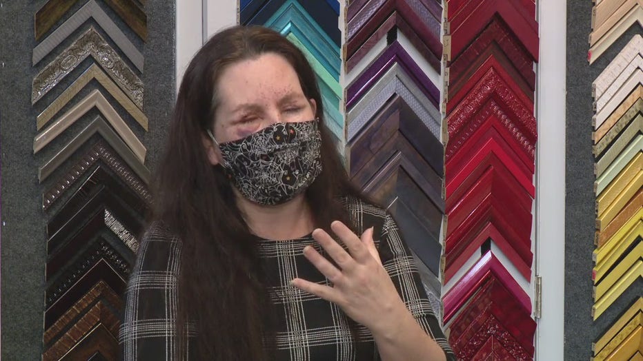 Cindy Eckley wears a cloth mask over her nose and face and stands in front of a frame shop, with damage to her eyes