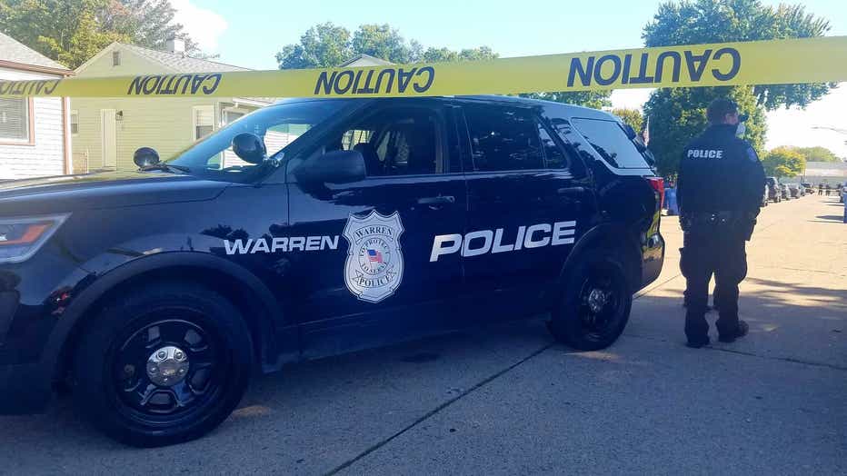 A Warren police cruiser is parked behind yellow caution tape, with an officer standing nearby.