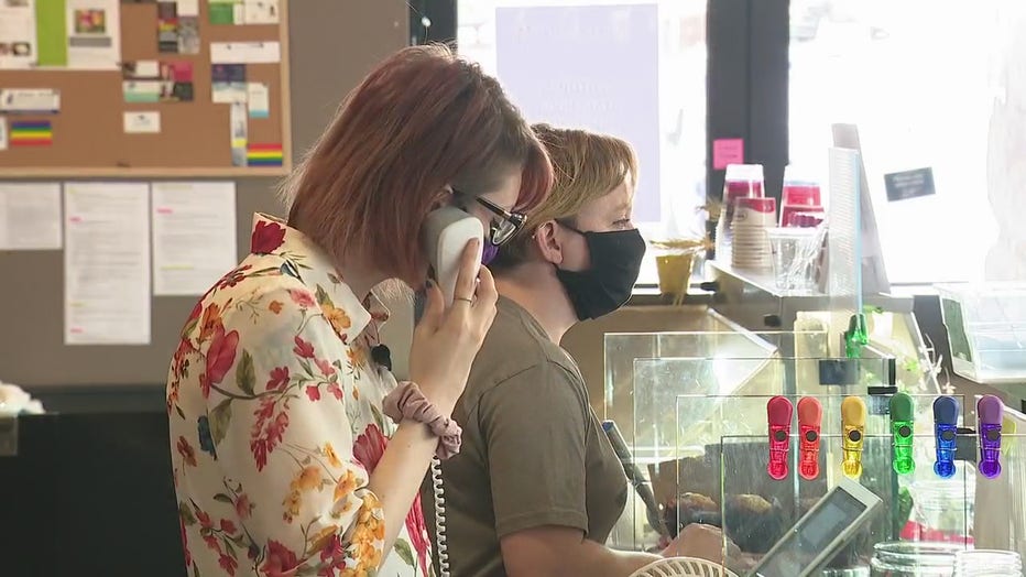Two women stand behind a restaurant counter, one on a phone call and the other speaking with a customer. 
