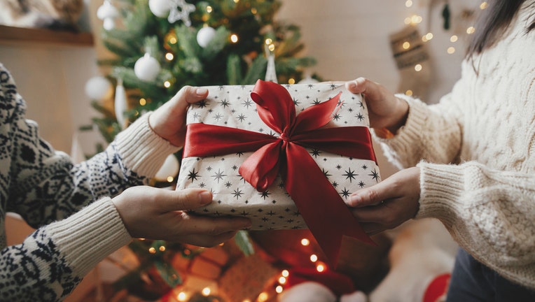 Couple exchanging christmas gift with red bow on background of christmas tree with lights. Stylish couple hands holding present with red ribbon close up in festive decorated room. Happy Holidays