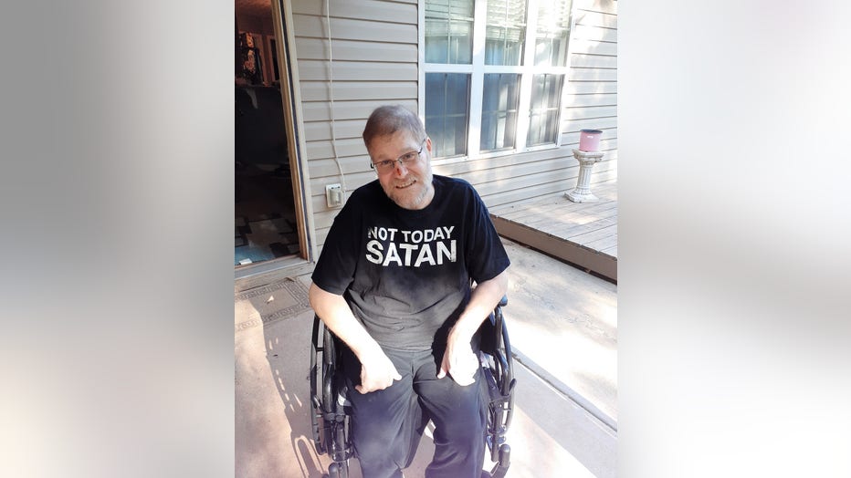 Man in wheelchair sits on front porch, smiling and wearing a t-shirt that reads 