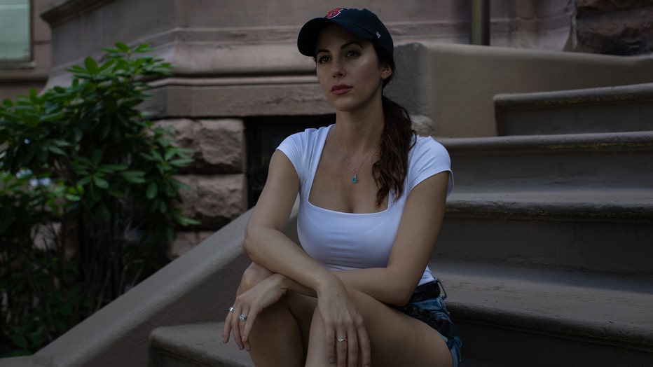 Woman sits on steps of building, wearing a baseball hat, with her arms crossed over her knees.