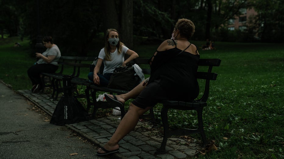 Woman sits on park bend, wearing a mask, talking with another woman, who is holding a cane. 