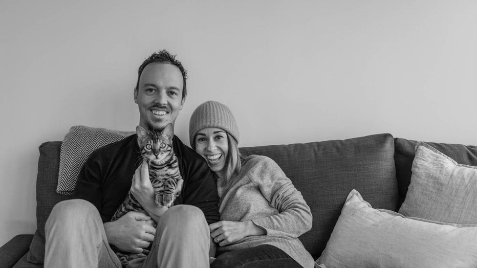 Couple sits on couch, smiling, holding a cat in their lap.