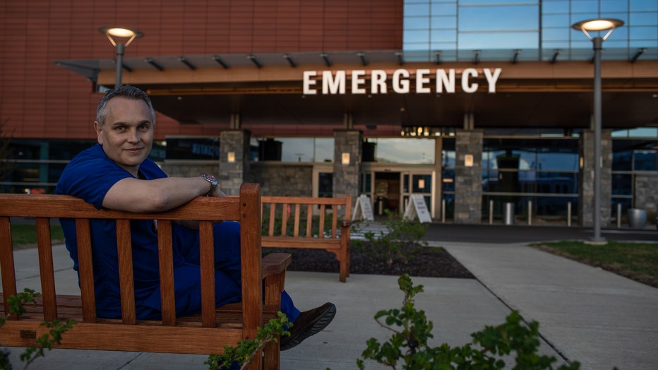 Doctor in scrubs sits outside in front of a hospital emergency sign.