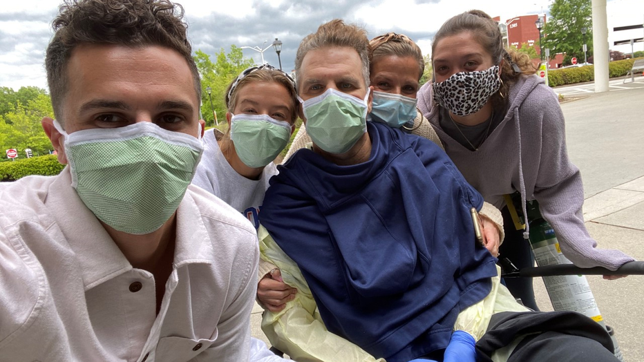 Man in wheelchair is surrounded by his family. All are wearing facemasks and he is wearing gloves.