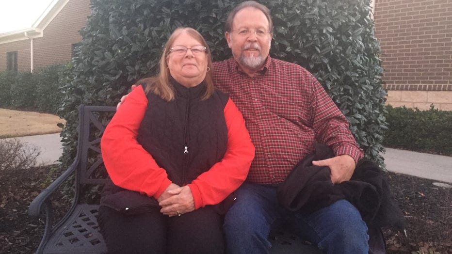 Woman and man pose while sitting on a bench. She has shoulder length hair and glasses. He has grey hair and a beard. They are smiling.