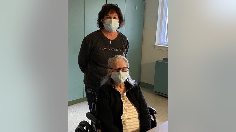 Woman in mask stands behind her elderly mother, who is in a wheelchair. 