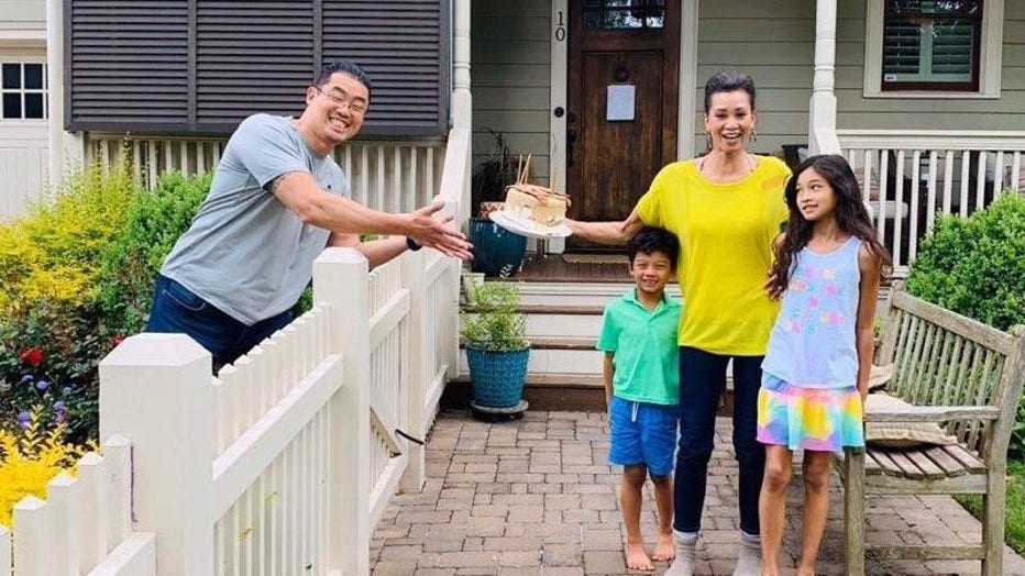 Man leans over fence, handing his wife a birthday cake. She is smiling and standing with their two young children.