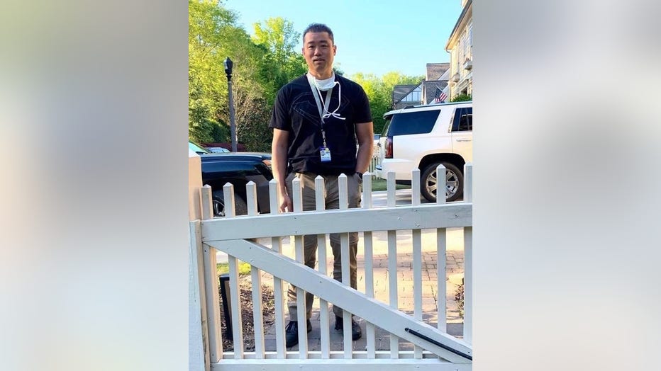 Man in hospital scrubs stands outside picket fence in front of his home.