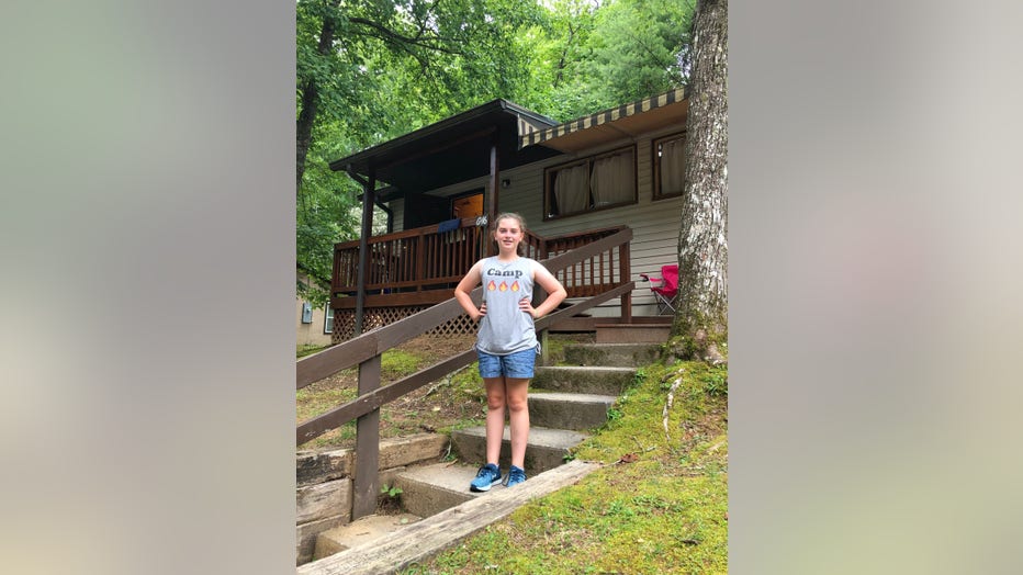 Young girls stands on steps outside her summer camp. She is wearing a t-shirt that says "camp" and is smiling, with her hands on her hips. She looks happy.
