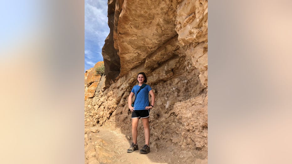 Young girl stands in front of rock formation with her hands on her hips. She is wearing shorts and a t-shirt and is smiling at the camera.