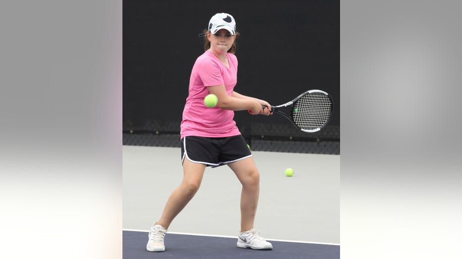 Girl in baseball hat swings a tennis racket at a ball.
