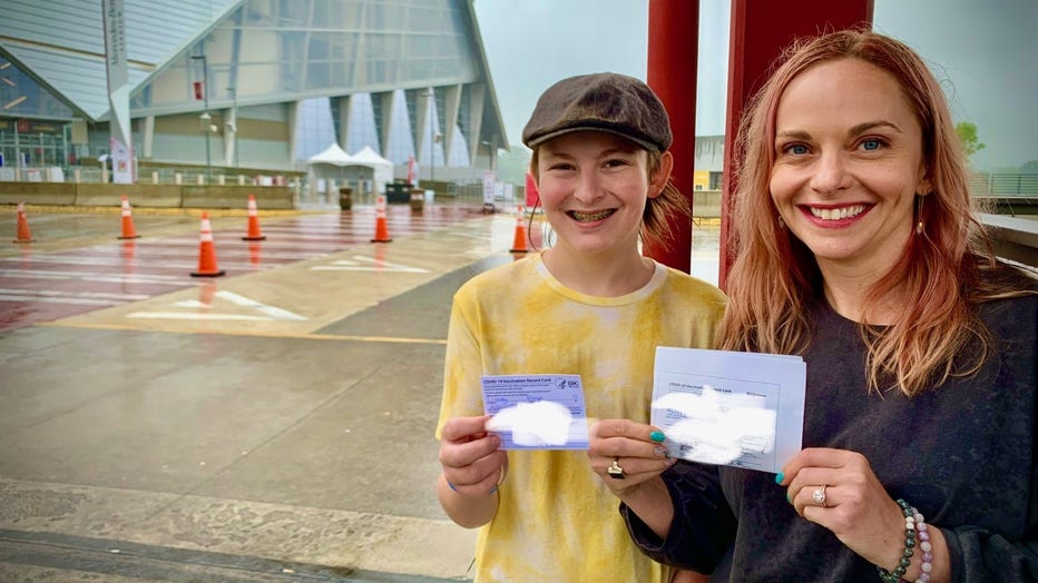 Teen boy wearing cap poses with his mother, both of them holding their COVID-19 vaccine cards, outside Atlanta's Mercedes-Benz Stadium.