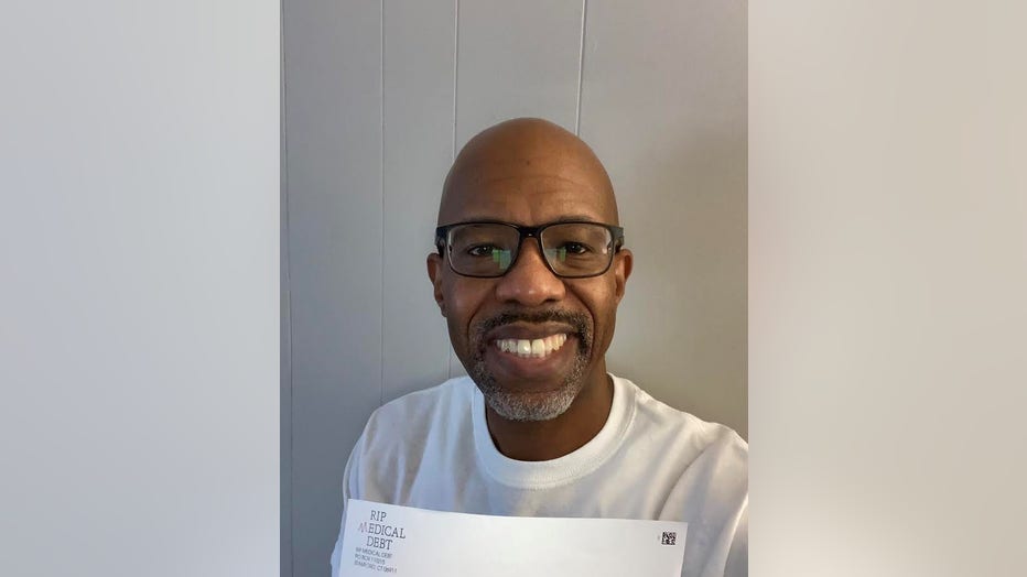 Smiling man with dark skin and shaved head holds a white envelope from RIP Medical Debt. 