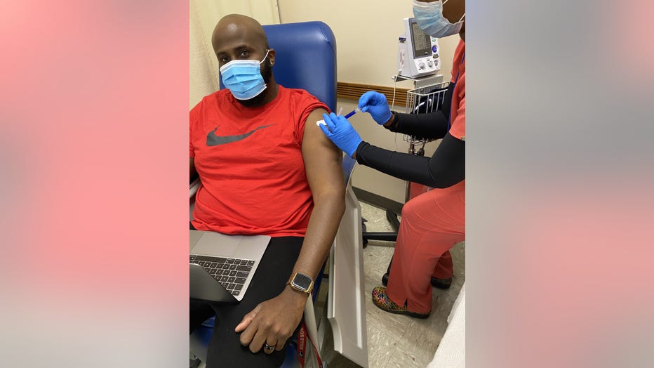 Black man with shaved head receives a shot from a nurse. He is wearing a mask and sitting in an exam room with a laptop computer in his lap.