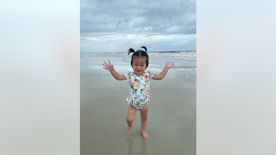 Little girl from China plays on the beach, her arms lifted in the air. She's smiling and wearing a toddler's bathing suit.