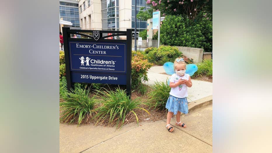 Blonde three-year-old girl wearing fairy wings stands in front of the Emory-Children's Center 