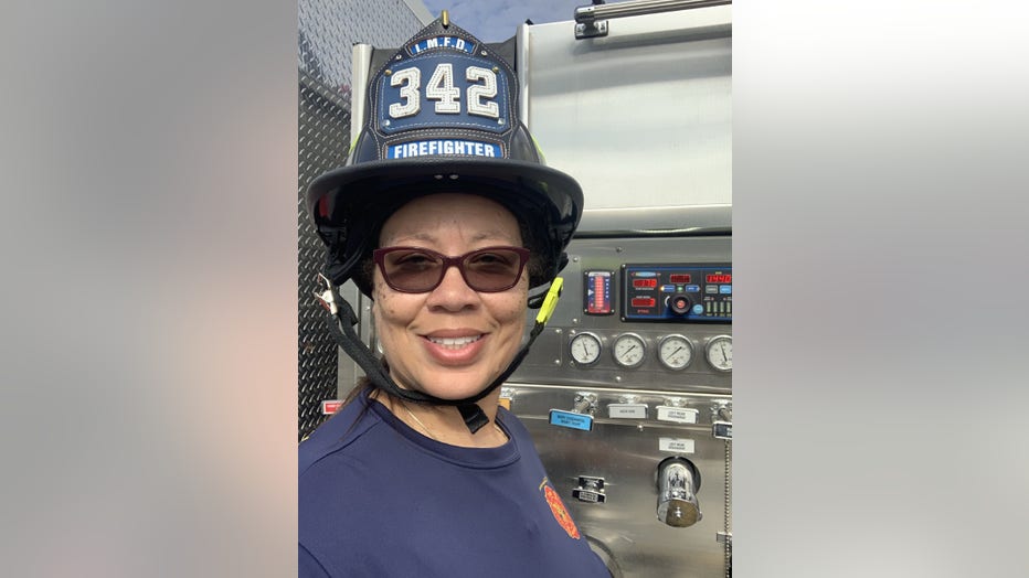 Black woman smiles. She's wearing a fire helmet, and is standing next to a fire rig.