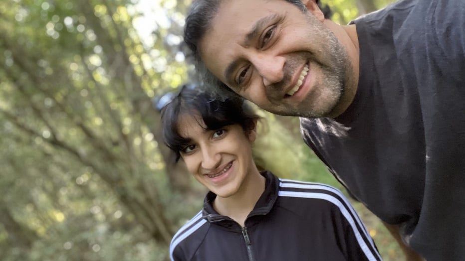 Father and daughter stand together in an outdoor wooded area. They are smiling at the camera, taking a selfie.