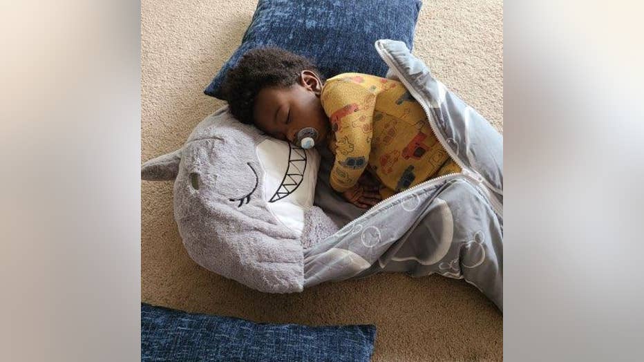 Young African American toddler sleeps on the floor, wrapped up in a shark sleeping bag. He has a pacifier in his mouth and is wearing pajamas.