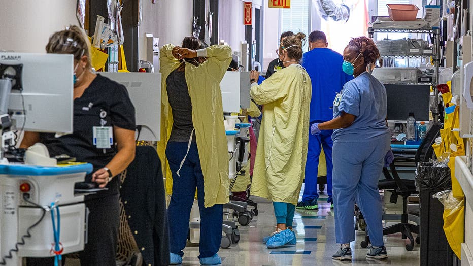 A crowded hospital hallway, where staff members wear protective equipment like masks and gowns