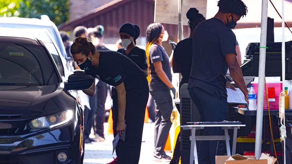 Workers in scrubs and protective clothing stand inside a testing and vaccination tent as drivers pull through a tented area.