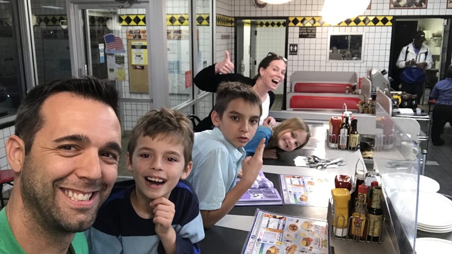 Family smiles and gestures at the camera while sitting at the counter of a Waffle House. The father is holding the camera and they have 3 children smiling between him and the mother, who is waving in the background.