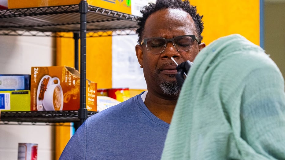 A Black man wearing glasses closes his eyes as a woman in a headscarf inserts a COVID-19 testing swab into his nose.