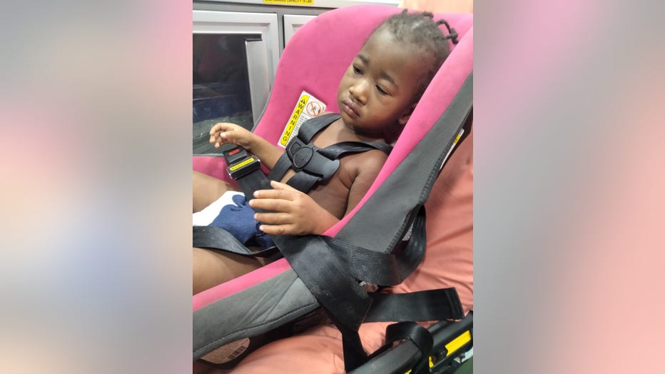 African American toddler sits in a car seat strapped to a gurney in the back of an ambulance.
