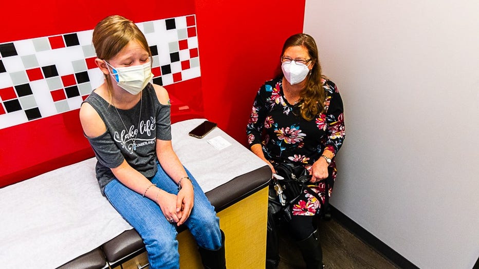 10-year-old girl sits on an exam table in her doctor's office awaiting a COVID-19 shot. Her mother is sitting in a chair in the corner of the room.