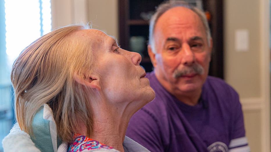 Man with grey hair and mustache looks at his wife whose faces is angled to the side and leaning into a chair. She is thin, five years after being diagnosed with early-onset Alzheimer's disease.