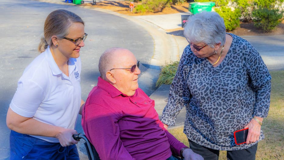 Woman pushed older man in wheelchair. outdoors. It is sunny and his wife is standing next to him talking to both of them.