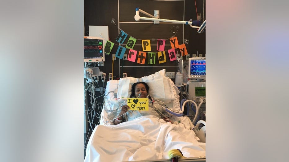 Pregnant woman poses in her ICU bed in front of a happy birthday sign. She is holding a sing that reads "I love you, love Mom."