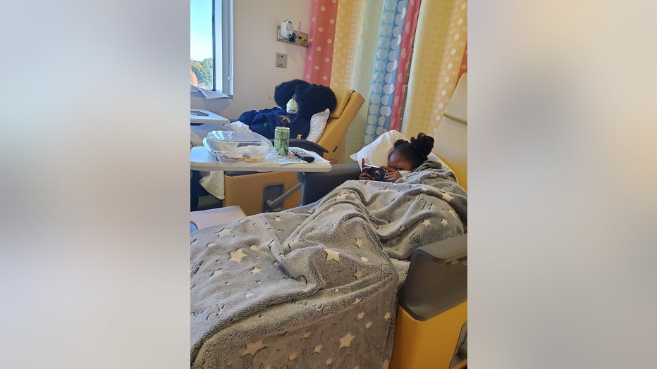 Two young girls sit in infusion chairs covered by blankets.