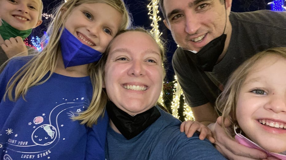 Family of 5 poses for a selfie in front of some holiday lights. They are all smiling and squeezing into the photo, while the mom smiles and holds the cellphone camera.