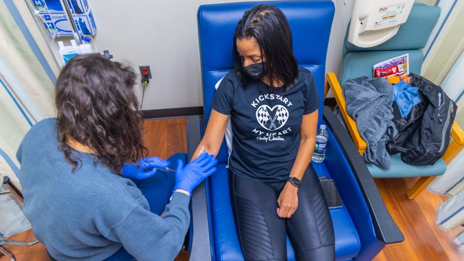 A woman sits in an infusion chair while a nurse places a needle in her arm to draw blood. 